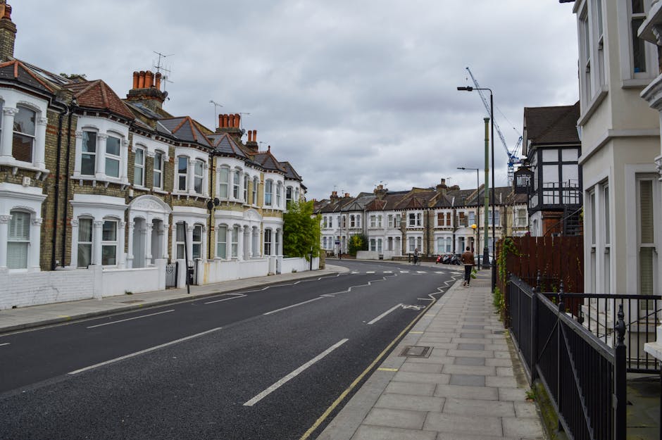 A quiet residential street in Dalston Kingsland featuring Victorian terraced houses with bay windows, decorative brickwork, and white-painted facades. The pavement on the right side is lined with a bl