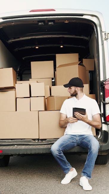 A young man with a beard, dressed in a white T-shirt, blue jeans, white sneakers, and a black cap, is sitting on the edge of an open moving van in an outdoor setting. The van’s cargo area is filled wi