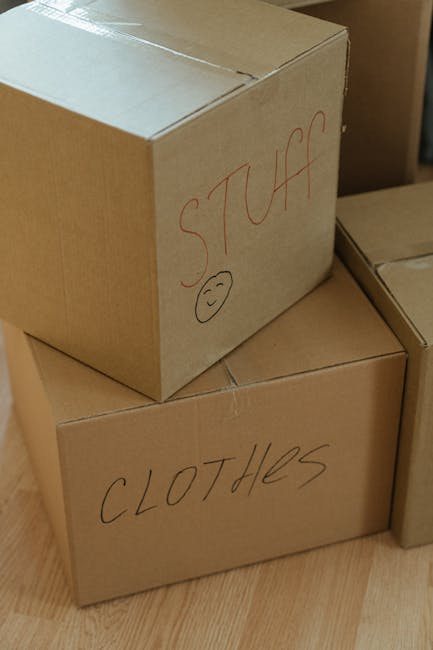 A young woman with curly brown hair tied in a bun, wearing a loose-fitting olive green top, is inside a home during a packing and moving process. She is leaning over a large cardboard box that is posi