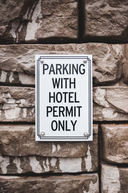 Four blue parking permit signs mounted on metal poles are positioned in a row on a paved surface in front of a modern building with a vertically ridged metal facade. The signs feature a white 'P' symb